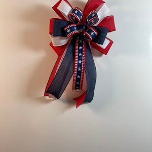 May include: A decorative bow featuring red, white, and blue ribbons. The bow has a layered design with a central section of blue ribbon adorned with white stars. Long, flowing tails extend from the bow, adding to its festive appearance.