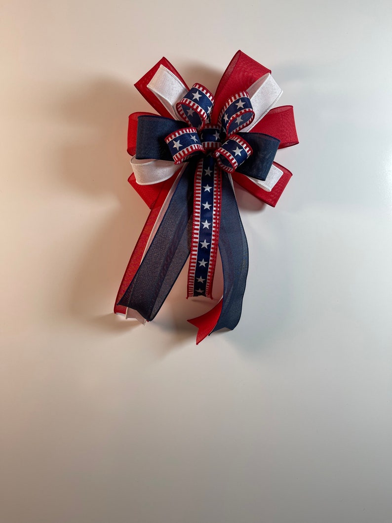 May include: A decorative bow featuring red, white, and blue ribbons. The bow has a layered design with a central section of blue ribbon adorned with white stars. Long, flowing tails extend from the bow, adding to its festive appearance.