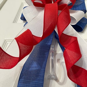 May include: A decorative bow featuring red, white, and blue ribbons. The bow is made of layered ribbons with a sheer texture, and is attached to a clear plastic clip. The color scheme is patriotic.