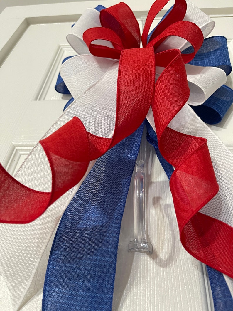 May include: A decorative bow featuring red, white, and blue ribbons. The bow is made of layered ribbons with a sheer texture, and is attached to a clear plastic clip. The color scheme is patriotic.