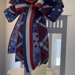 Puede incluir: Una linterna blanca est&aacute; adornada con un gran lazo decorativo. El lazo presenta cintas rojas, blancas y azules con lunares y patrones de cuadros. Una vela blanca est&aacute; dentro de la linterna, creando un tema patri&oacute;tico.