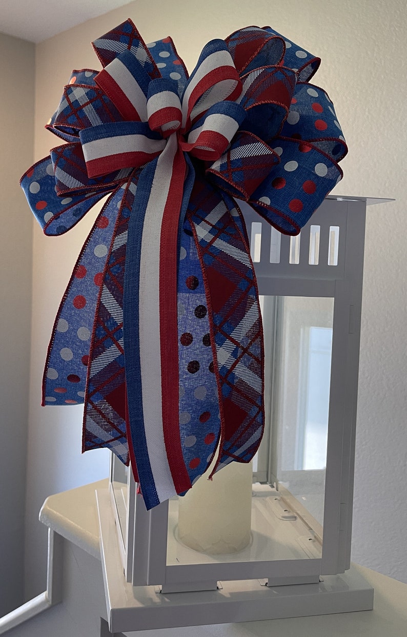Puede incluir: Una linterna blanca est&aacute; adornada con un gran lazo decorativo. El lazo presenta cintas rojas, blancas y azules con lunares y patrones de cuadros. Una vela blanca est&aacute; dentro de la linterna, creando un tema patri&oacute;tico.