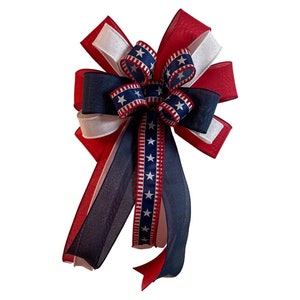 May include: A decorative bow in red, white, and blue, featuring a patriotic design. The bow has multiple layers of ribbon, with a central section adorned with stars. The long streamers add a festive touch, perfect for Independence Day or other patriotic events.