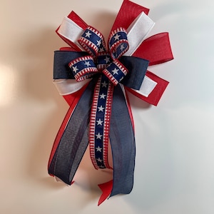 May include: A decorative bow in red, white, and blue, featuring patriotic star and stripe patterns. The bow is made of layered ribbons, with a central knot and long, flowing tails. Perfect for Independence Day or other patriotic celebrations.