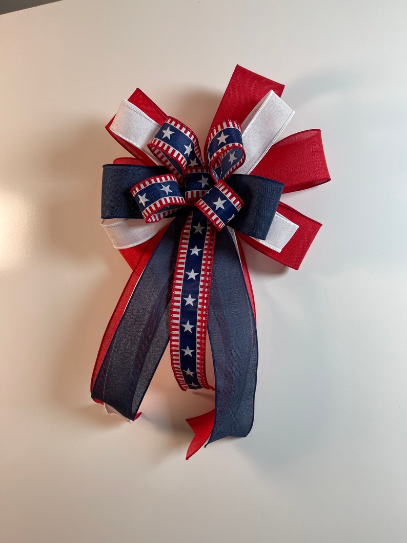 May include: A decorative bow in red, white, and blue, featuring patriotic star and stripe patterns. The bow is made of layered ribbons, with a central knot and long, flowing tails. Perfect for Independence Day or other patriotic celebrations.