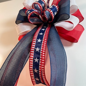 May include: A patriotic bow featuring red, white, and blue ribbons. The bow has a layered design with a central knot and long, flowing streamers. The ribbons display star patterns and stripes, creating a festive and decorative accent.