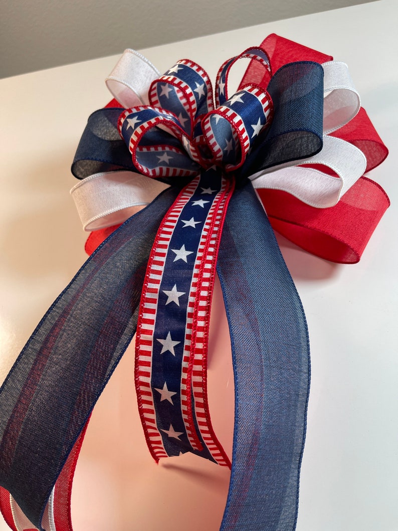 May include: A patriotic bow featuring red, white, and blue ribbons. The bow has a layered design with a central knot and long, flowing streamers. The ribbons display star patterns and stripes, creating a festive and decorative accent.