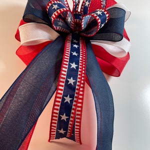 May include: A patriotic bow featuring red, white, and blue ribbons. The bow has a layered design with a central cluster and long, flowing tails. The ribbons display stars and stripes, perfect for Independence Day or other patriotic events.