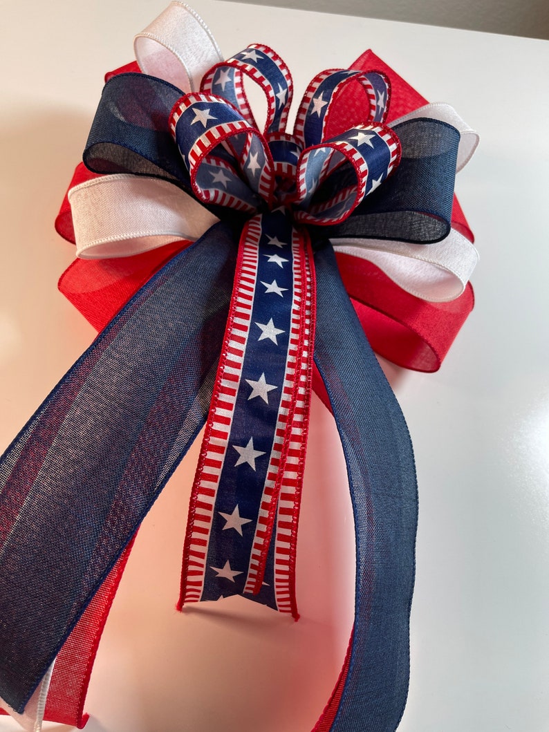 May include: A patriotic bow featuring red, white, and blue ribbons. The bow has a layered design with a central cluster and long, flowing tails. The ribbons display stars and stripes, perfect for Independence Day or other patriotic events.