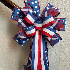 Puede incluir: Un lazo decorativo con colores y patrones patri&oacute;ticos. El lazo tiene cintas azules con lunares blancos, rayas rojas y blancas y un dise&ntilde;o de cuadros. El lazo est&aacute; sujeto a una barandilla de madera.