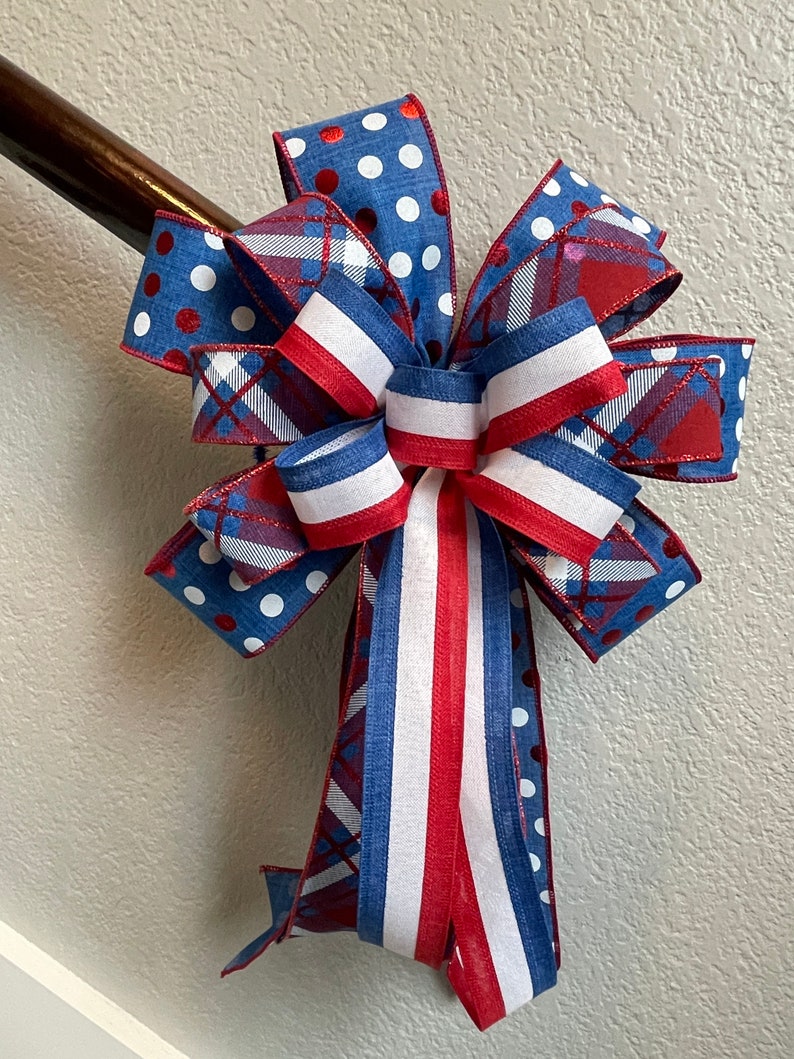 Puede incluir: Un lazo decorativo con colores y patrones patri&oacute;ticos. El lazo tiene cintas azules con lunares blancos, rayas rojas y blancas y un dise&ntilde;o de cuadros. El lazo est&aacute; sujeto a una barandilla de madera.