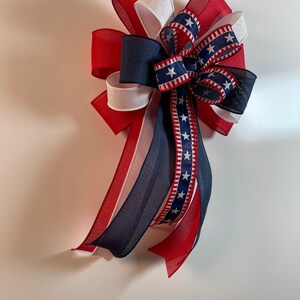 May include: A patriotic bow featuring red, white, and blue ribbons. The bow has a layered design with a central section of navy blue ribbon adorned with white stars. The ribbons are arranged to create a decorative accent.