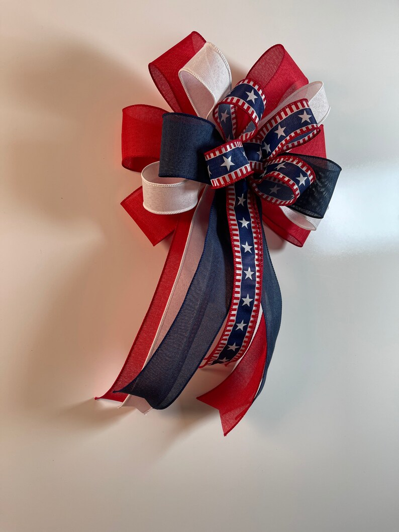 May include: A patriotic bow featuring red, white, and blue ribbons. The bow has a layered design with a central section of navy blue ribbon adorned with white stars. The ribbons are arranged to create a decorative accent.