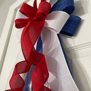 May include: A patriotic door bow featuring red, white, and blue ribbons. The bow has a layered design with a clear plastic ring for hanging. The red ribbon is sheer and curled, adding a decorative touch to the festive bow.