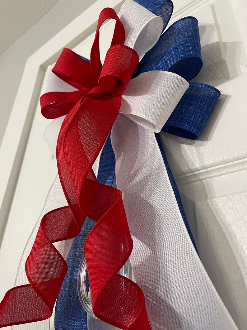 May include: A patriotic door bow featuring red, white, and blue ribbons. The bow has a layered design with a clear plastic ring for hanging. The red ribbon is sheer and curled, adding a decorative touch to the festive bow.