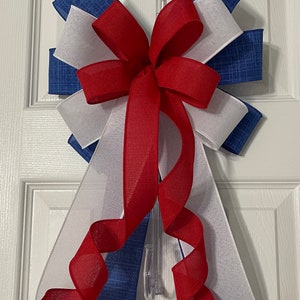 May include: A decorative bow in red, white, and blue, likely for patriotic or holiday decor. The bow features layers of fabric and ribbon, with a central red bow and cascading red ribbon. The bow is attached to a clear plastic hanger.