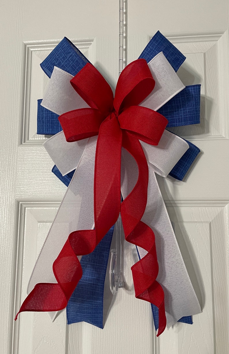 May include: A decorative bow in red, white, and blue, likely for patriotic or holiday decor. The bow features layers of fabric and ribbon, with a central red bow and cascading red ribbon. The bow is attached to a clear plastic hanger.