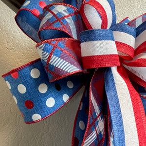 Puede incluir: Un lazo decorativo con colores patri&oacute;ticos. El lazo tiene cintas azules con lunares blancos y detalles rojos. Otras cintas tienen un estampado de cuadros en rojo, blanco y azul. El lazo est&aacute; atado con una cinta a rayas rojas y blancas.