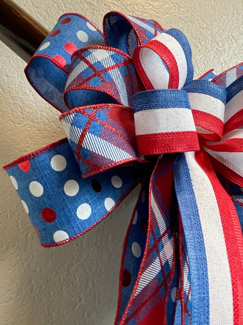 Puede incluir: Un lazo decorativo con colores patri&oacute;ticos. El lazo tiene cintas azules con lunares blancos y detalles rojos. Otras cintas tienen un estampado de cuadros en rojo, blanco y azul. El lazo est&aacute; atado con una cinta a rayas rojas y blancas.