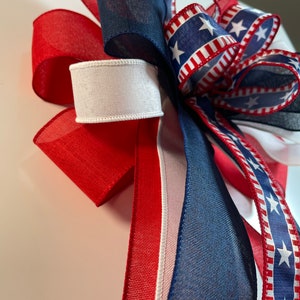 May include: A patriotic bow featuring red, white, and blue ribbons. The bow includes a ribbon with white stars on a blue background, and solid red, white, and navy blue ribbons. The bow is likely for a wreath or other decorative use.