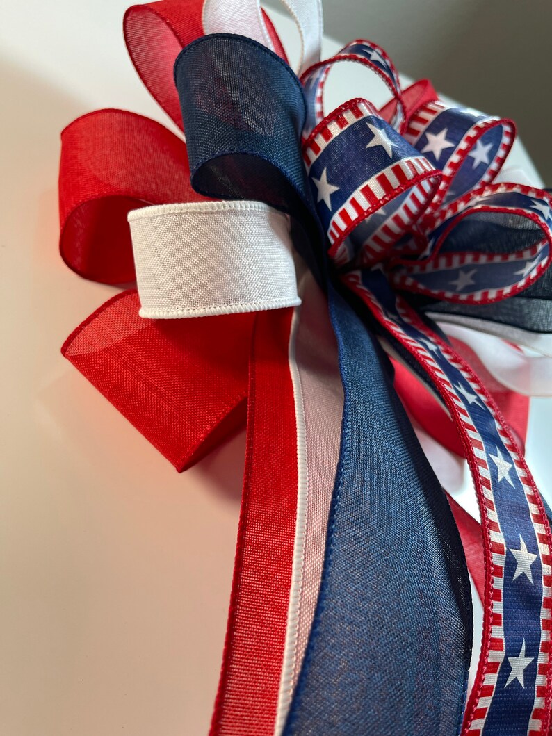 May include: A patriotic bow featuring red, white, and blue ribbons. The bow includes a ribbon with white stars on a blue background, and solid red, white, and navy blue ribbons. The bow is likely for a wreath or other decorative use.