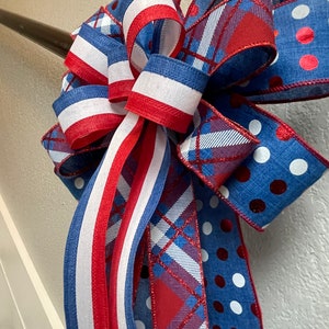 Puede incluir: Un lazo decorativo con colores patri&oacute;ticos. El lazo tiene cintas azules, blancas y rojas con lunares, rayas y patrones de cuadros. El lazo probablemente se utiliza para decoraci&oacute;n, posiblemente para una festividad o evento especial.