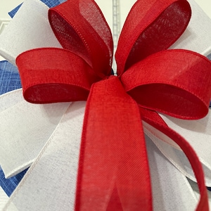 May include: Close-up of a decorative bow featuring red, white, and blue ribbons. The bow is made of layered fabric, with the red ribbon forming the prominent central element. The white and blue ribbons are visible in the background.