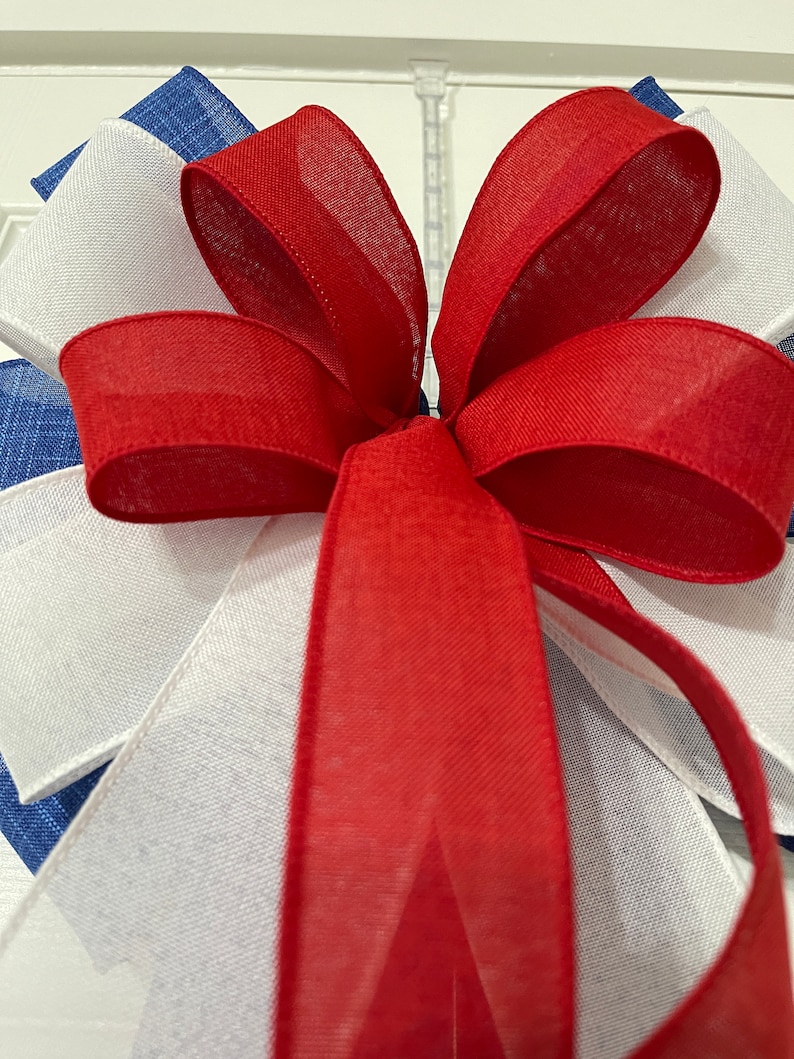 May include: Close-up of a decorative bow featuring red, white, and blue ribbons. The bow is made of layered fabric, with the red ribbon forming the prominent central element. The white and blue ribbons are visible in the background.