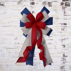 May include: A decorative bow featuring red, white, and blue ribbons. The bow has a layered design with a central red bow and cascading ribbons in shades of white, blue, and red. The bow is likely for festive or patriotic decorations.