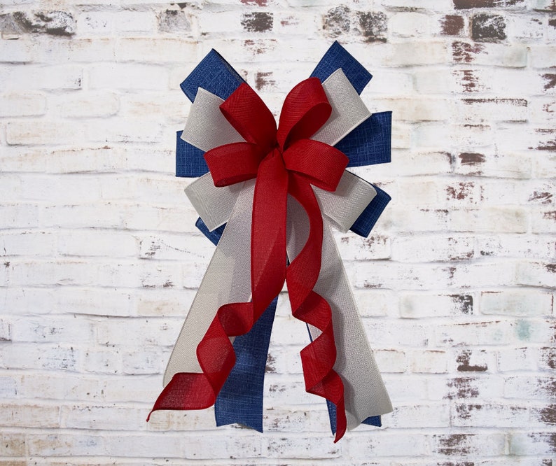 May include: A decorative bow featuring red, white, and blue ribbons. The bow has a layered design with a central red bow and cascading ribbons in shades of white, blue, and red. The bow is likely for festive or patriotic decorations.