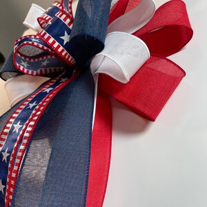 May include: A decorative bow featuring patriotic colors. The bow is made of red, white, and blue ribbons. Some ribbons have a pattern of white stars on a blue background, while others are solid colors. The bow is likely for festive decorations.