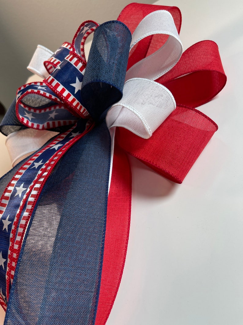 May include: A decorative bow featuring patriotic colors. The bow is made of red, white, and blue ribbons. Some ribbons have a pattern of white stars on a blue background, while others are solid colors. The bow is likely for festive decorations.