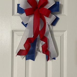 May include: A decorative bow in red, white, and blue, hanging on a white door. The bow features layers of blue fabric with a textured pattern, white fabric, and a red sheer ribbon. The bow is attached to a clear plastic hanger.