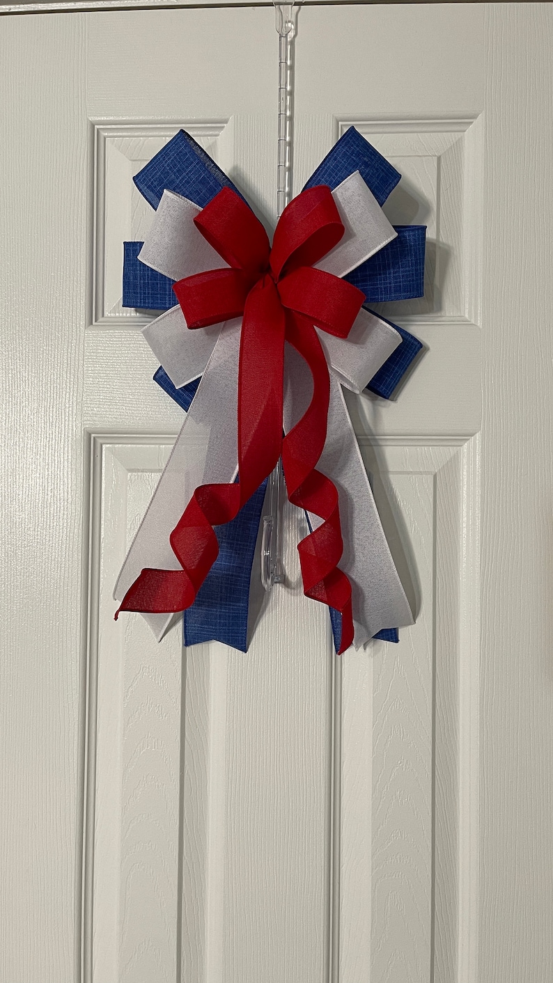 May include: A decorative bow in red, white, and blue, hanging on a white door. The bow features layers of blue fabric with a textured pattern, white fabric, and a red sheer ribbon. The bow is attached to a clear plastic hanger.