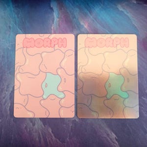 May include: Two rectangular cards with the word "MORPH" in pink block letters at the top. The cards have a repeating pattern of abstract shapes in pink and light blue. One card has a holographic finish.