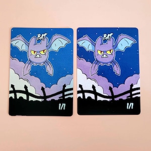 May include: Two illustrated cards depicting a purple bat with yellow eyes and wings against a blue night sky with clouds. The word "BAT" is above the bat. A black fence is at the bottom, with the number "1/1" below.