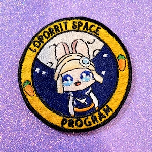 Peut inclure: Un patch brodé jaune et bleu avec un lapin de dessin animé portant un casque et une combinaison spatiale bleue et blanche. Le patch porte le texte "LOOPERIT SPACE PROGRAM" autour du bord.