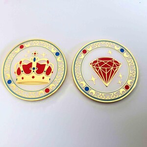 May include: Two gold-coloured round pins. One features a red, gold, and blue crown design. The other pin has a red diamond design with gold stars. Both pins have a white background and a gold border with blue and red accents.