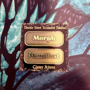 May include: Two double-sided keyword tokens with the words "Morph" and "Manifest" in gold lettering. The tokens are on a black card with the text "Double Sided Keyword Tokens" and "Clover Alters". The background is a teal and purple pattern.