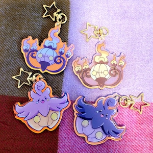 May include: Four colorful acrylic keychains featuring cartoon-style characters. Two keychains depict a flame-shaped design with a pumpkin-like face, while the other two showcase a character with a flower-like design. Each keychain has a gold star charm.