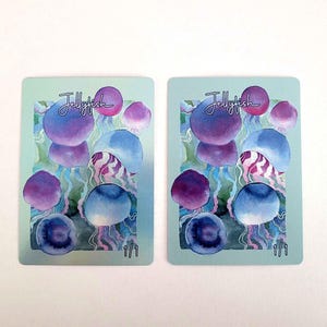May include: Two rectangular cards featuring a watercolour illustration of jellyfish. The jellyfish are in shades of blue, purple, and pink, with the word "Jellyfish" in cursive. The cards have a light teal background.