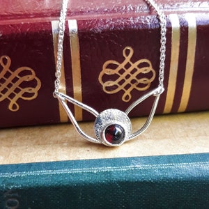 May include: A silver necklace with a red gemstone pendant. The pendant is set in a silver circle with a textured surface. The necklace is on a red book with gold lettering.