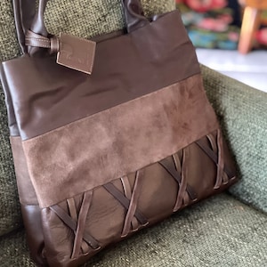 May include: A brown leather tote bag with a suede panel and a braided leather design. The bag has a top handle and a tag that says "2N1".