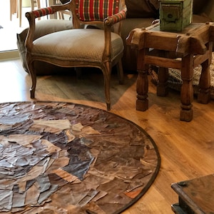 Könnte beinhalten: Ein runder, brauner Lederteppich mit Patchwork-Muster. Der Teppich besteht aus verschiedenen Lederstücken, die zusammengenäht sind. Der Teppich befindet sich in einem Wohnzimmer mit einem Holzstuhl und einem Holztisch.