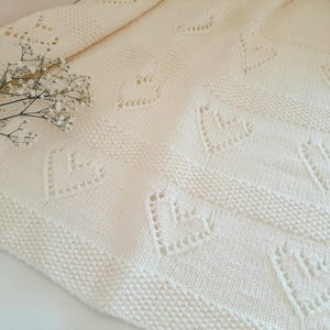 Peut inclure: Couverture en tricot crème avec des découpes en forme de cœur. La couverture présente un motif texturé et une bordure décorative. Une brindille de fleurs blanches repose sur la couverture. Idéale pour la décoration intérieure.