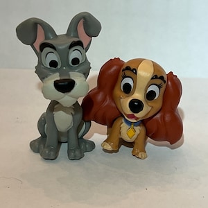 May include: Two figurines of cartoon dogs, one gray and one brown. The gray dog has large ears and a mischievous expression. The brown dog has long, flowing hair and a blue collar with a yellow tag. Both are smiling.