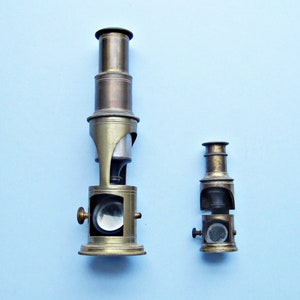 May include: Two antique brass microscopes, one larger than the other, on a light blue background. The larger microscope has a round base and a tall, cylindrical body with a lens at the top. The smaller microscope has a shorter body and a smaller lens. Both microscopes are in good condition and have a vintage look.
