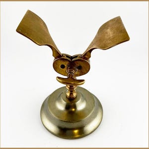 Puede incluir: Un objeto de latón vintage con una base circular, un vástago central y dos elementos en forma de ala curvados hacia arriba. La pieza tiene una apariencia mecánica detallada, con un pequeño mango y juntas intrincadas. El latón tiene una pátina cálida y envejecida.