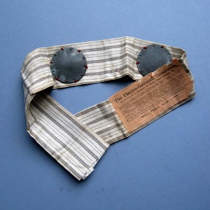 Puede incluir: Un cinturón antiguo de generadores electrogalvánicos. El cinturón está hecho de tela a rayas con dos placas metálicas circulares adjuntas. Una etiqueta de papel con texto está adjunta al cinturón. La etiqueta dice "The Electro-Galvanic Generators".