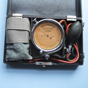 Puede incluir: Un maletín médico negro abierto que contiene un oscilotonómetro vintage con una esfera beige y un bisel plateado. El kit también incluye un manguito de tela gris y una pera negra con tubos rojos. El dial indica "OSCILLOTONOMETRE".